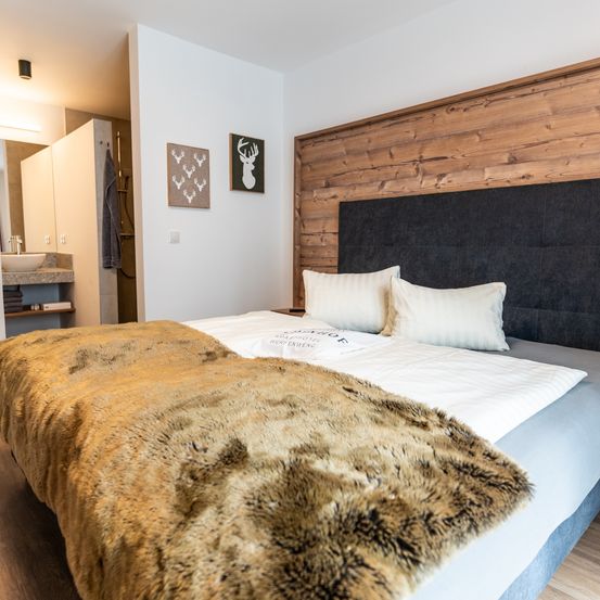 Ein Schlafzimmer mit einem Bett, einem hölzernen Kopfteil, zwei Kissen und einer Pelzdecke. Links befindet sich ein Waschbecken und ein Handtuch, sowie zwei Bilderrahmen an der Wand.