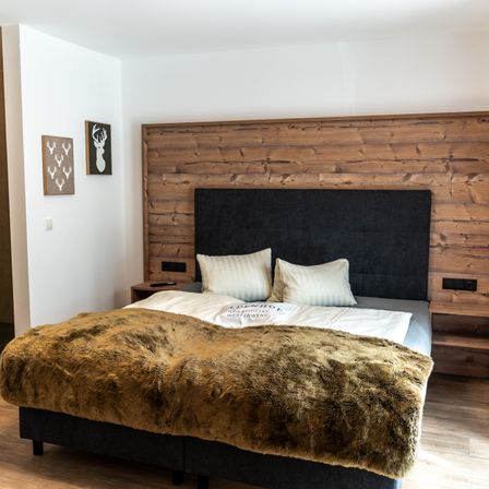 Ein Schlafzimmer mit einem hölzernen Kopfteil zeigt ein Bett, das mit einer braunen Pelzdecke bedeckt ist, zwei weißen Kissen und zwei gerahmten Bildern an der Wand.