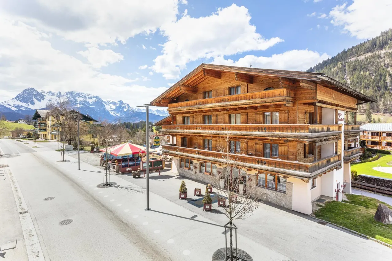 Ein großes Holzgebäude mit vielen Fenstern und Balkonen steht an einer Bergstraße, mit einem Zelt und Bäumen in der Nähe.
