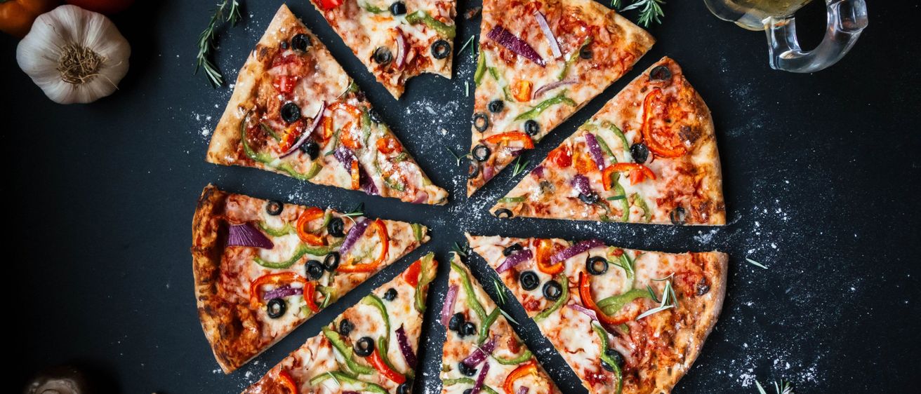 Ein Nahaufnahme einer Reihe von Pizzastücken mit Toppings wie roten und grünen Paprika, schwarzen Oliven und Rosmarin auf einer dunklen Oberfläche.