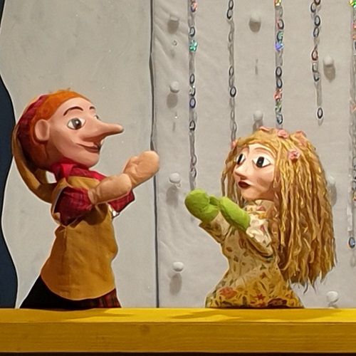 Zwei Puppen, eine mit einer langen Nase und die andere mit blonden Haaren, stehen auf einer Bühne vor einem weißen Hintergrund. Die Puppe mit der langen Nase trägt ein gelbes Hemd und ein rotes Halstuch, während die andere ein Blumenkleid und grüne Handschuhe trägt.