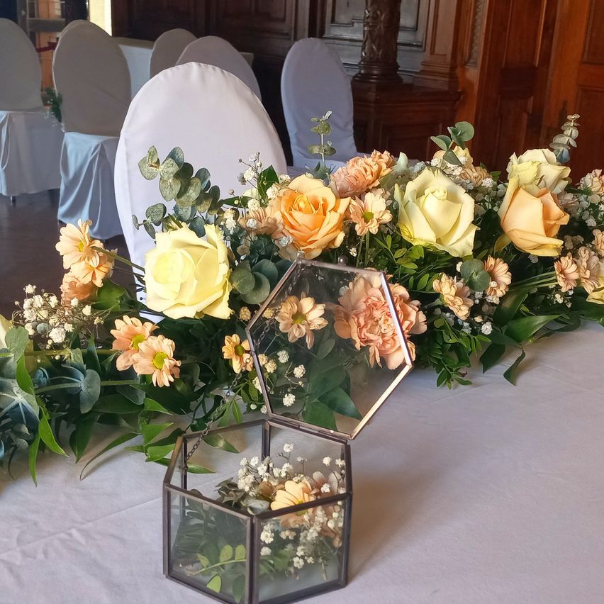 Ein Tisch ist mit einer langen Blumendekoration geschmückt, die einen sechseckigen Glasbehälter enthält. Die Blumen bestehen aus Rosen und Gänseblümchen in Pfirsichtönen und Creme. Stühle sind im Hintergrund ordentlich angeordnet.
