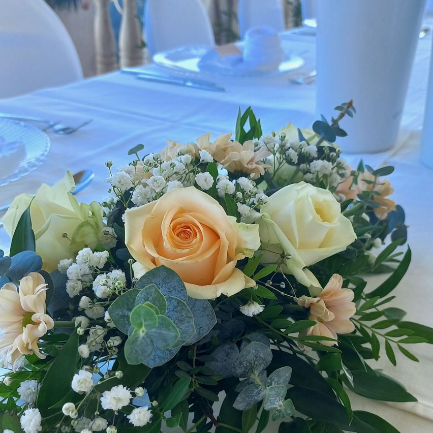 Eine Tischdekoration mit einer Blumenarrangement in der Mitte, bestehend aus Rosen, Gänseblümchen und Grün auf einer weißen Tischdecke.
