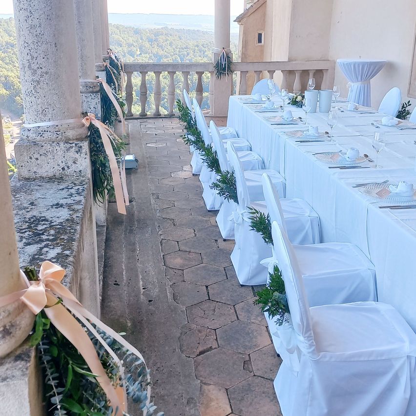 Ein langer Tisch ist mit weißen Tischdecken und Stühlen auf einer Terrasse im Freien mit Blick auf das Meer und die Berge gedeckt.