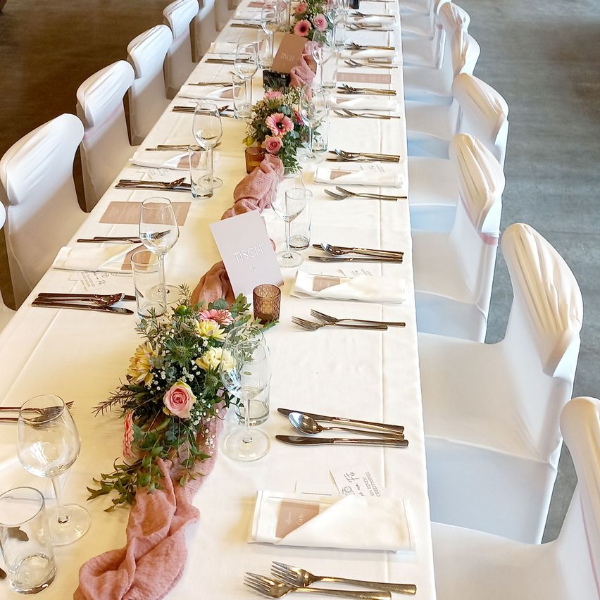 Ein langer Tisch mit weißen Stühlen ist für eine Dinnerparty vorbereitet. Der Tisch hat weiße Tischdecken, Weingläser und Besteck. Eine Blumenarrangement mit rosa und gelben Blumen ist auf dem Tisch platziert.