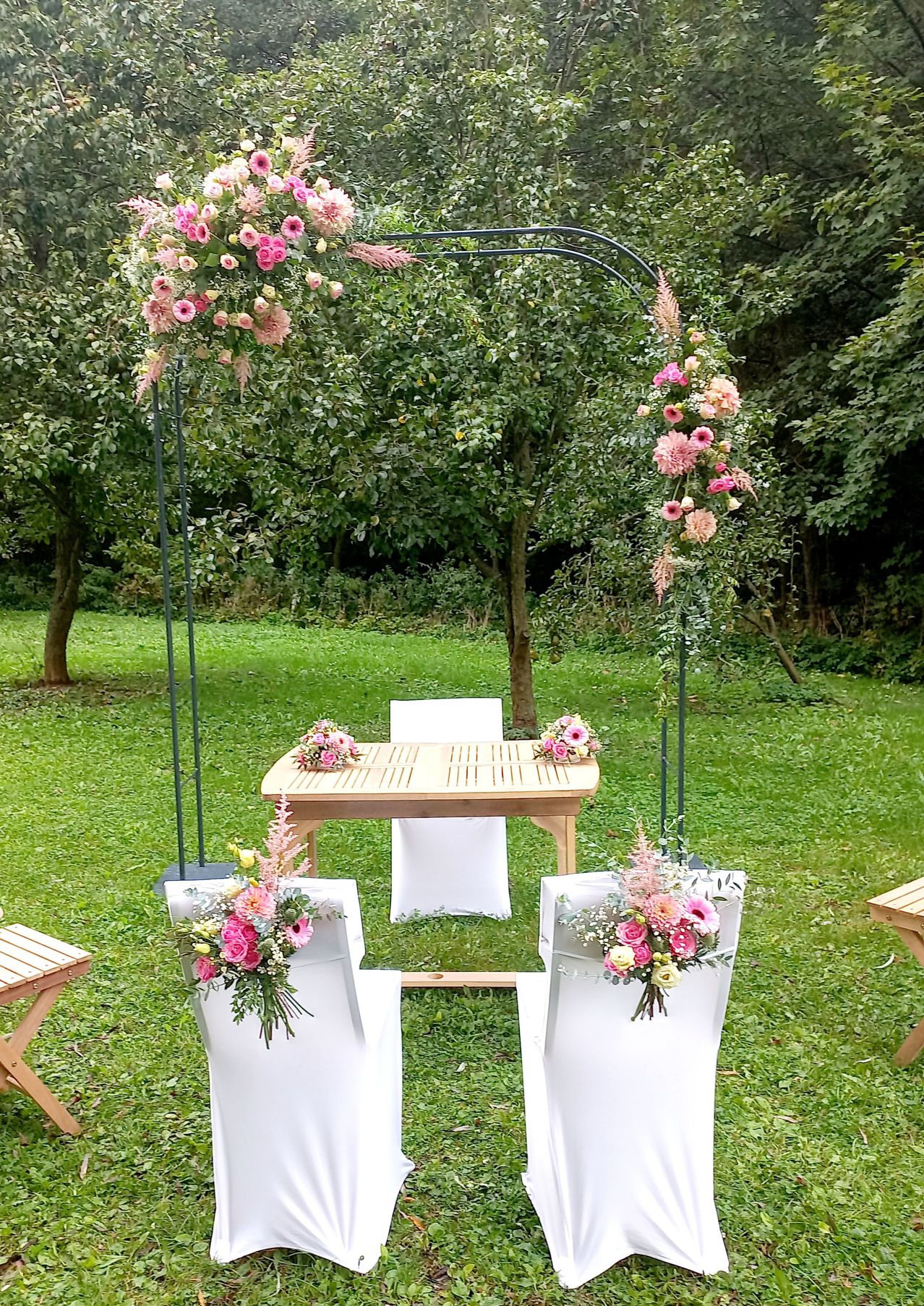 Ein floraler Bogen mit rosa Blumen schmückt eine Hochzeitsdekoration auf einem Rasen, mit einem Holztisch und Stühlen darunter. Grüne Bäume und Büsche sind im Hintergrund.