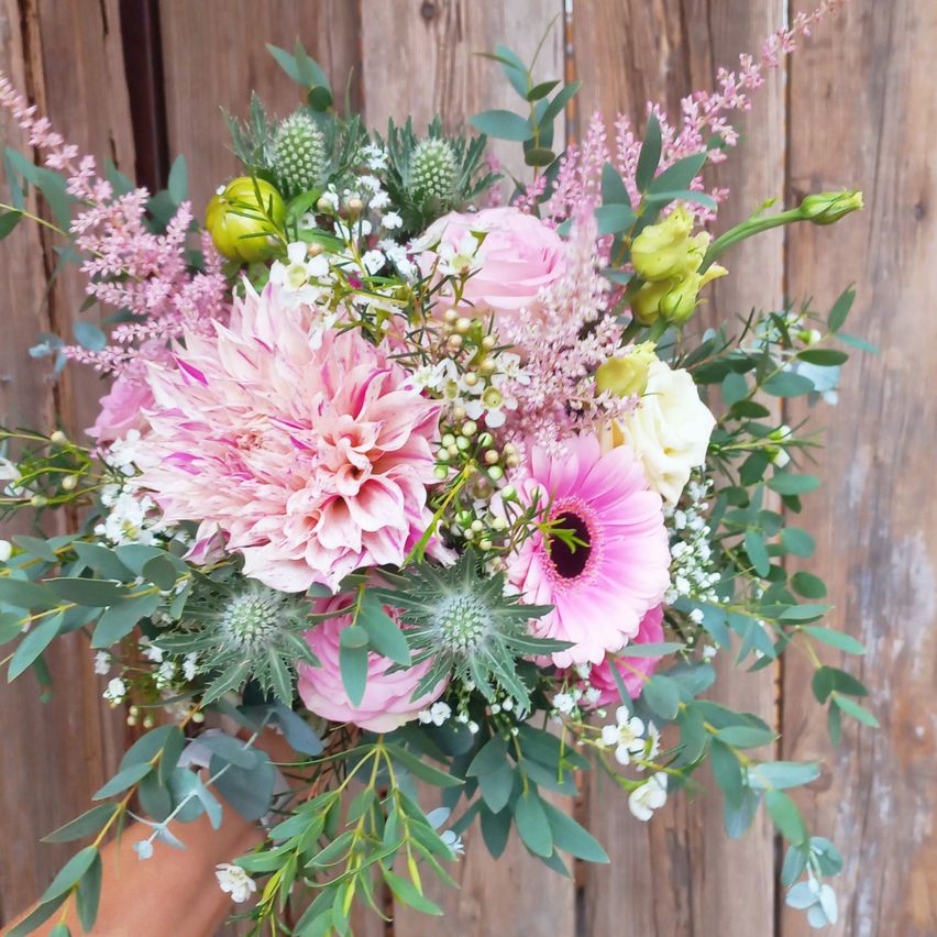 Ein Blumenstrauß mit rosa und weißen Blumen, einschließlich Dahlien, Rosen und Babys Atem, wird von einer Hand vor einem hölzernen Hintergrund gehalten.