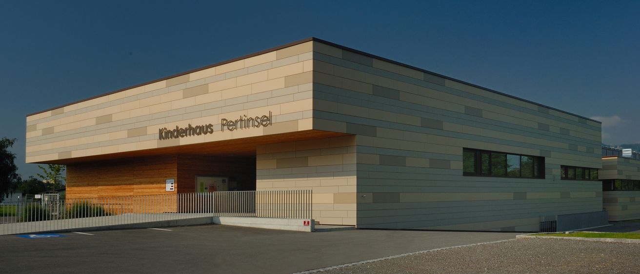 Die Vorderseite eines Gebäudes mit beigefarbener Wand und Holzeingang, beschriftet mit 'Kinderhaus Pertinsel', unter einem klaren blauen Himmel.