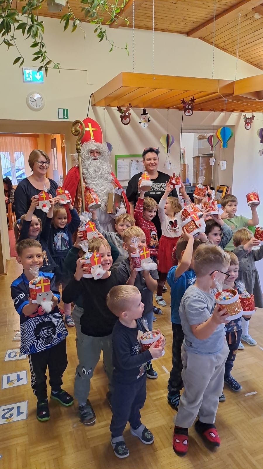 Mehrere Kinder halten dekorierte Körbe mit Leckereien darin hoch. Ein Mann, der als Weihnachtsmann verkleidet ist, steht hinter ihnen und lächelt. Zwei Frauen, eine mit Brille, sind anwesend. Ballons und Dekorationen hängen von der Decke.