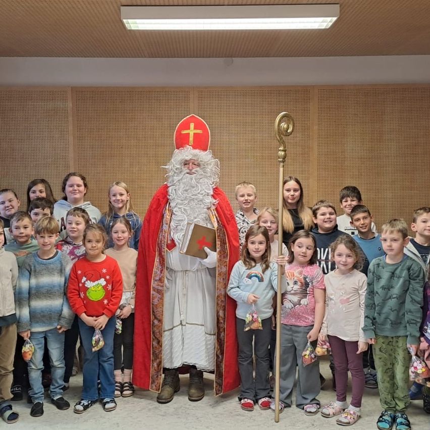 Eine Gruppe von Kindern steht um einen Mann, der als Weihnachtsmann verkleidet ist. Er hält ein Buch und einen Stab, und einige Kinder halten Süßigkeiten. Sie alle scheinen für ein Foto zu posieren.