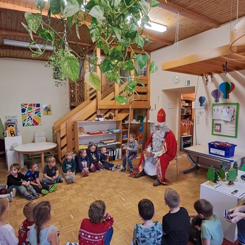 Eine Gruppe von Kindern sitzt auf dem Boden in einem Klassenzimmer. Ein Mann, der als Weihnachtsmann verkleidet ist, sitzt auf einem Stuhl in der Mitte des Raumes. Es gibt einen Tisch mit weißer Tischplatte und ein Regal mit Büchern.