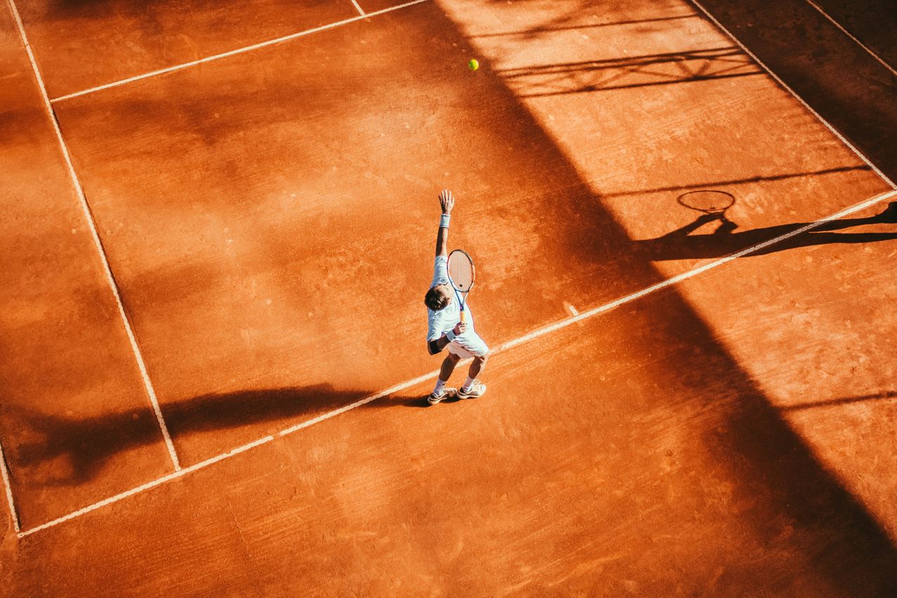 Ein Tennisspieler in einem blauen Shirt serviert einen gelben Tennisball auf einem Sandplatz bei hellem Sonnenlicht.