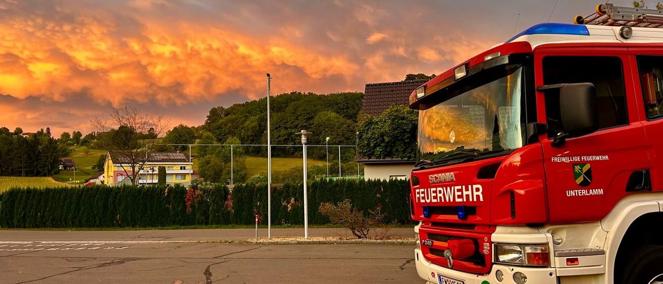 Ein roter Feuerwehrwagen steht auf einem Parkplatz mit einem Sonnenuntergang im Hintergrund.