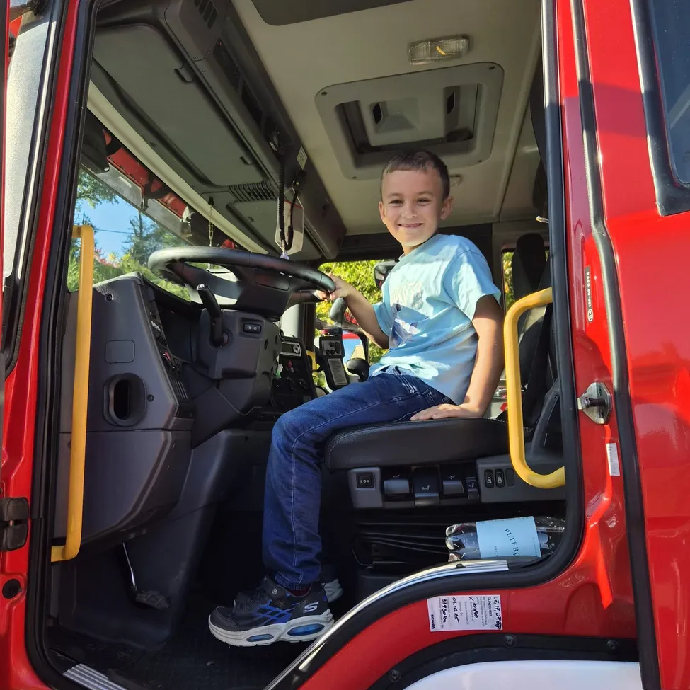Ein junger Junge sitzt im Fahrersitz eines roten Feuerwehrautos. Er lächelt und hält das Lenkrad.