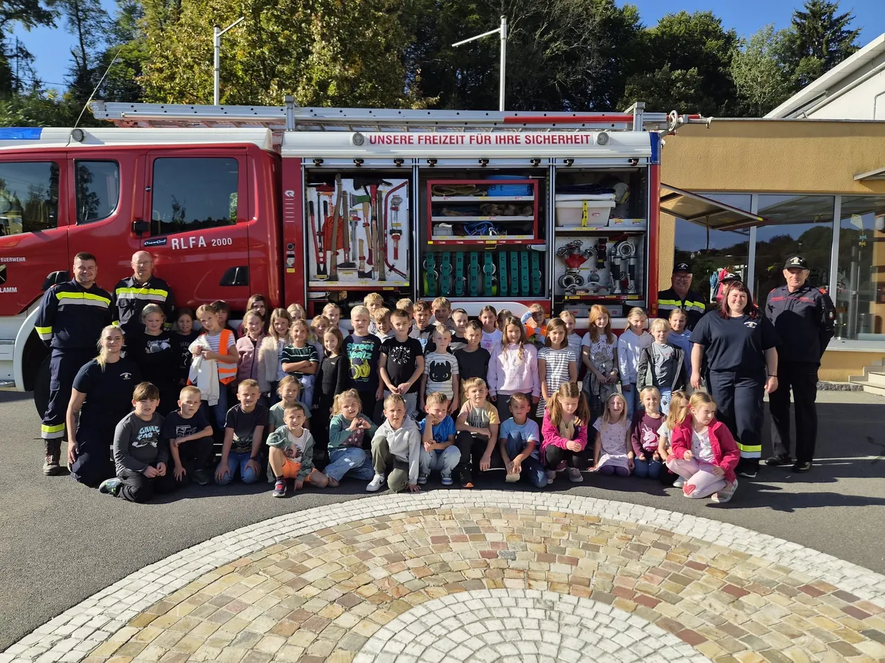 Eine Gruppe von Kindern und Erwachsenen posiert für ein Foto vor einem Feuerwehrauto. Das Feuerwehrauto ist rot und hat die Aufschrift 'Unsere Freiheit für Ihre Sicherheit'.