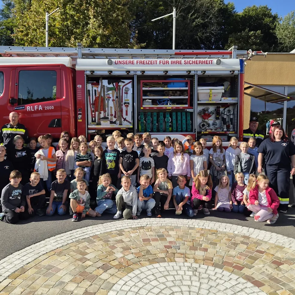 Eine Gruppe von Kindern und Erwachsenen posiert für ein Foto vor einem Feuerwehrauto. Das Feuerwehrauto ist rot und hat die Aufschrift 'Unsere Freiheit für Ihre Sicherheit'.