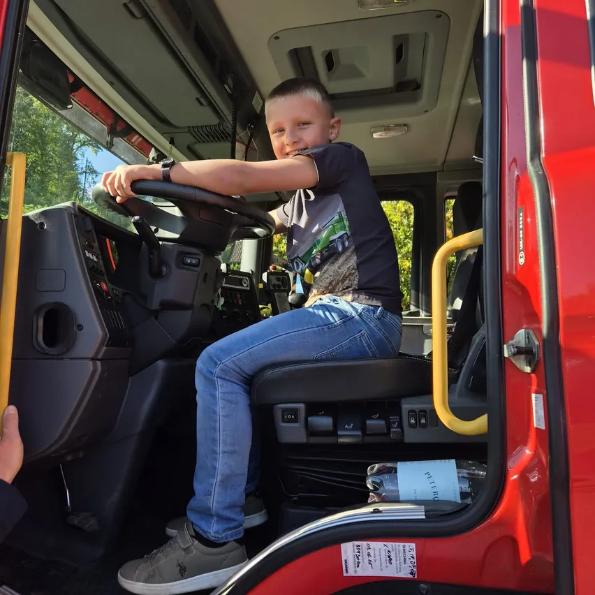 Ein junger Junge sitzt im Fahrersitz eines roten Feuerwehrwagens und hält das Lenkrad mit einem Lächeln im Gesicht.