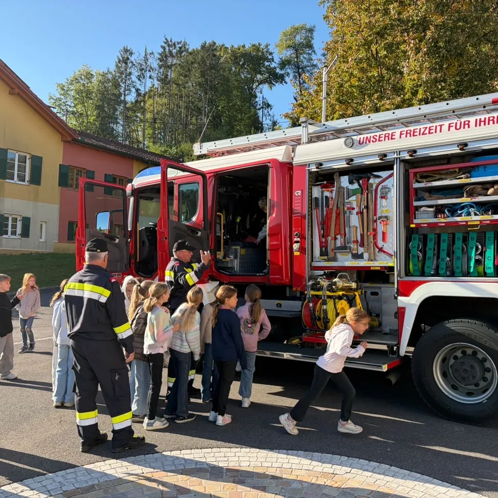 Eine Gruppe von Kindern ist um einen Feuerwehrwagen versammelt. Zwei Feuerwehrleute stehen neben dem Wagen, von denen einer auf etwas im Inneren des Wagens zeigt. Der Wagen ist vor einem Gebäude mit roter Wand und gelbem Dach geparkt.