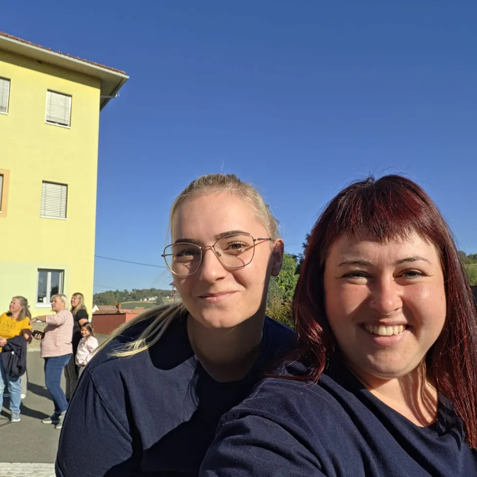 Zwei Frauen lächeln und posieren für ein Foto vor einem gelben Gebäude.