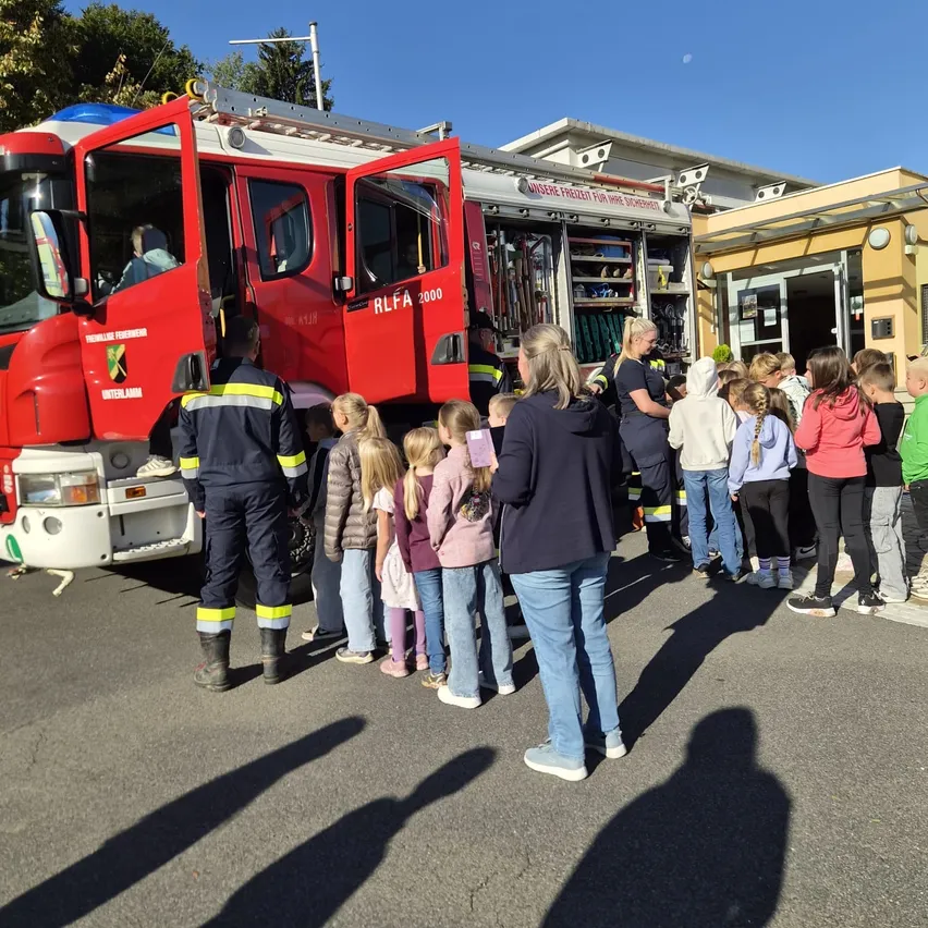 Eine Gruppe von Kindern und Erwachsenen steht um einen roten Feuerwehrwagen. Ein Feuerwehrmann steht neben dem Wagen.