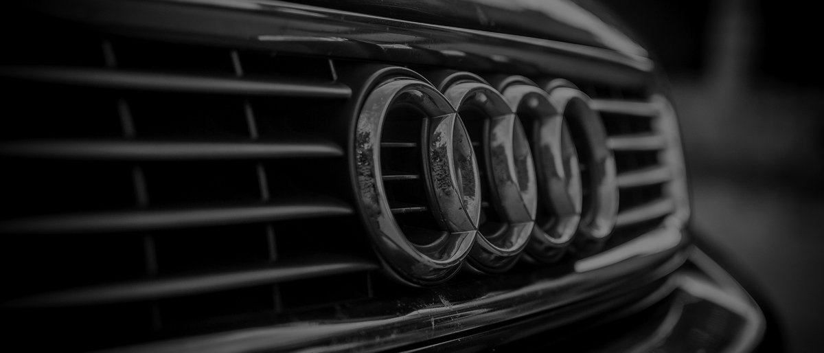 Schwarz-Weiß-Bild der Kühlergrill eines Audi-Autos mit dem Markenlogo. Das Logo besteht aus vier ineinandergreifenden Ringen, und der Kühlergrill hat ein Gittermuster.