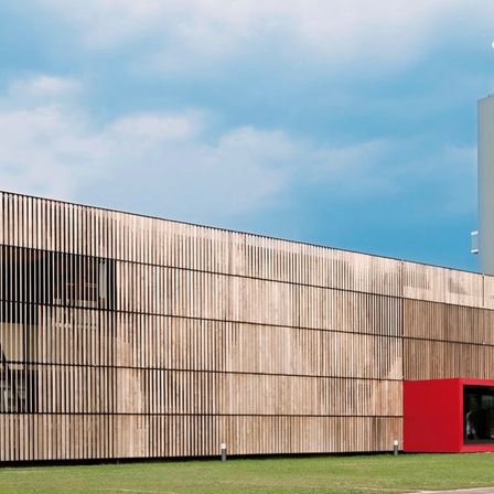 Außenansicht eines modernen Gebäudes mit einer großen Holzfassade, die vertikale Lamellen aufweist. Rechts befindet sich eine kleine rote Struktur, und der Himmel ist teilweise bewölkt.