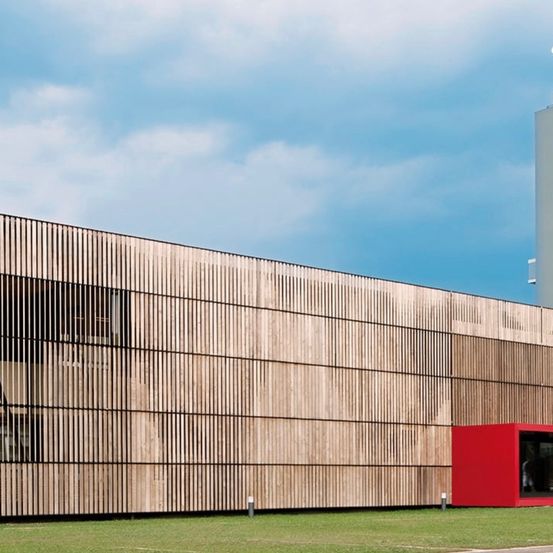 Außenansicht eines modernen Gebäudes mit einer großen Holzfassade, die vertikale Lamellen aufweist. Rechts befindet sich eine kleine rote Struktur, und der Himmel ist teilweise bewölkt.