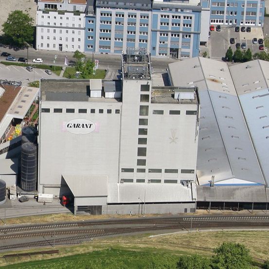 Luftaufnahme eines Industriegebäudes mit der Aufschrift Garant. Es ist groß mit mehreren Fenstern und an andere Gebäude angeschlossen. Es sind mehrere Autos in der Nähe geparkt.