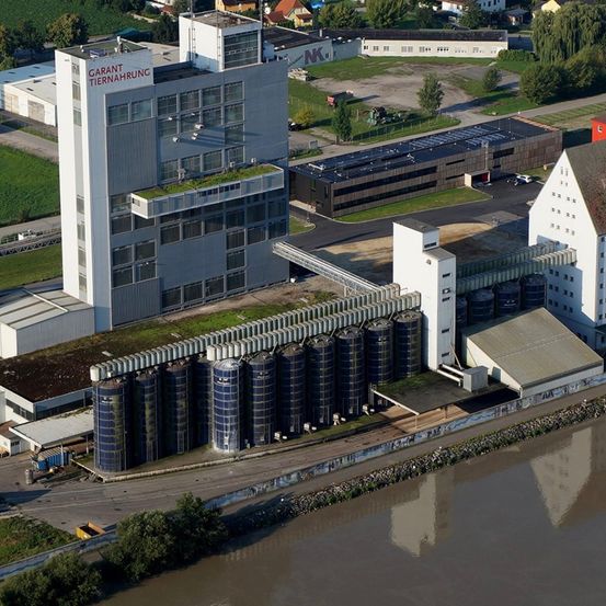 Luftaufnahme einer großen Industrieanlage mit einem markanten weißen Gebäude, das mit 'Garant Tiernahrung' beschriftet ist. In der Nähe befinden sich zahlreiche zylindrische Lagertanks, ein kleineres Gebäude und eine asphaltierte Straße mit einem Auto. Das umliegende Gebiet umfasst Bäume, Gras und weitere Gebäude.