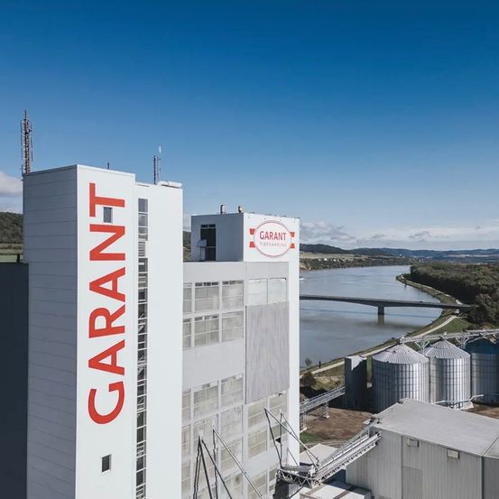 Ein Blick von oben auf ein Garant-Werk, das auf einem Gebäude groß geschrieben steht. In der Nähe fließt ein Fluss mit einer Brücke im Hintergrund vorbei.
