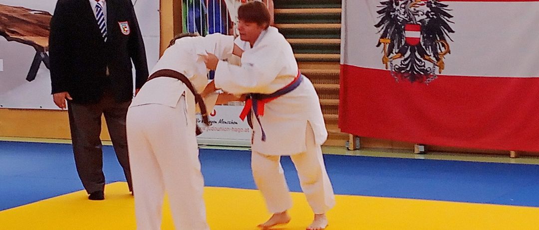 Zwei Personen üben Judo auf einem gelben Mattenbereich. Ein Mann in einem schwarzen Anzug steht in der Nähe. Der Text 'Save the date! 18. Oktober 2025' und 'Offene Österreichische Meisterschaft: Adapted Judo' steht auf der Matte. Eine Flagge ist hinter den Teilnehmern positioniert.