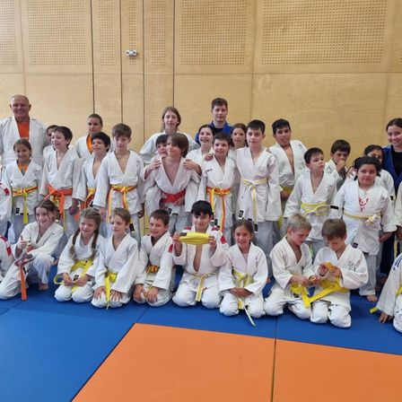 Eine Gruppe junger Karateschüler, Jungen und Mädchen, in weißen Uniformen und farbigen Gürteln, posiert für ein Foto in einer Turnhalle.