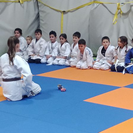 Eine Gruppe junger Kinder in weißen Kampfsportuniformen sitzt auf einem blau-orangefarbenen Teppich, wahrscheinlich in einem Kampfsportkurs.
