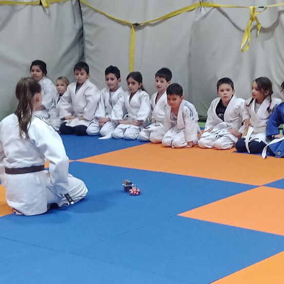 Eine Gruppe junger Kinder in weißen Kampfsportuniformen sitzt auf einem blau-orangefarbenen Teppich, wahrscheinlich in einem Kampfsportkurs.