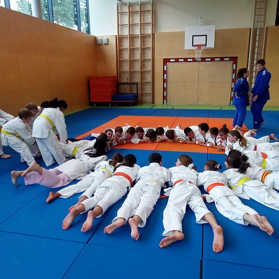 Gruppe junger Schüler trainiert Kampfsport in einer Turnhalle. Zwei Trainer stehen in der Nähe des Basketballkorbs.