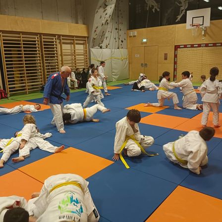 Mehrere Kinder üben Kampfsport auf einer blau-orangefarbenen Matte unter Aufsicht eines Erwachsenen.