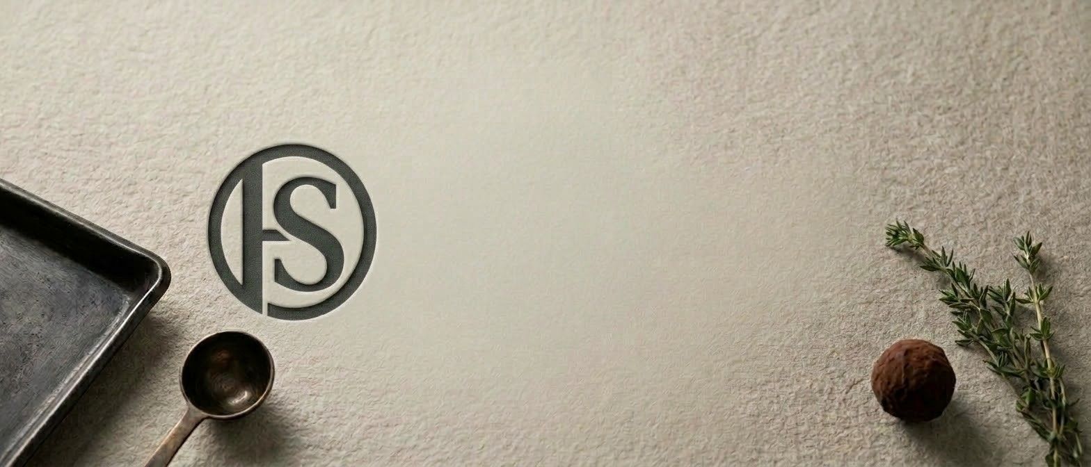 Eine weiße Oberfläche mit einem Logo, einem Löffel, einem Tablett, einem Rosmarinzweig und einer Haselnuss.
