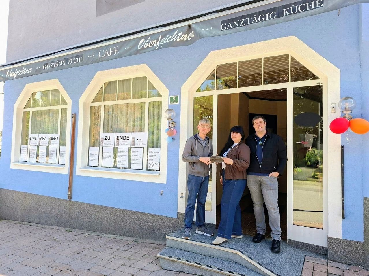 Drei Personen stehen auf den Stufen eines blauen Gebäudes mit einem Schild, das 'Ganztagig Kuche' besagt. Der Mann auf der linken Seite hält ein kleines Objekt, möglicherweise einen Schlüssel, und die Frau in der Mitte hält ein Stück Papier.