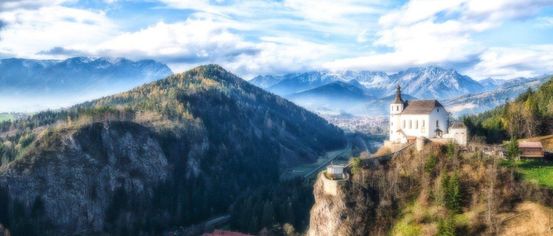 Eine weiße Kirche steht auf einem Felsen mit Blick auf ein Tal mit einer Straße und Gebäuden, umgeben von einem Wald und Bergen unter einem blauen Himmel mit Wolken.
