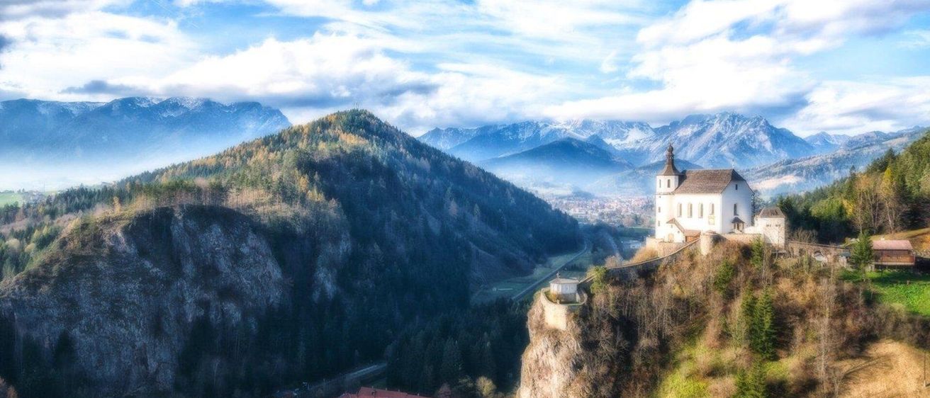 Eine weiße Kirche steht auf einem Felsen mit Blick auf ein Tal mit einer Straße und Gebäuden, umgeben von einem Wald und Bergen unter einem blauen Himmel mit Wolken.