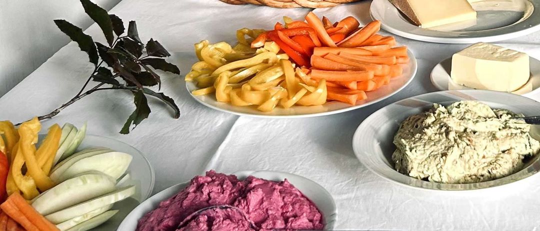 Ein Tisch ist mit verschiedenen Speisen gedeckt. Ein Korb mit Brot, verschiedene Käsesorten und geschnittenes Gemüse. Schalen mit Hummus und Rote-Bete-Dip. Frische Kräuter zur Seite.