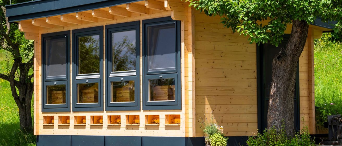 Ein kleines Holzhaus mit schwarzem Rand und mehreren Fenstern steht auf einem gepflasterten Gehweg, umgeben von üppigen grünen Bäumen und Pflanzen.