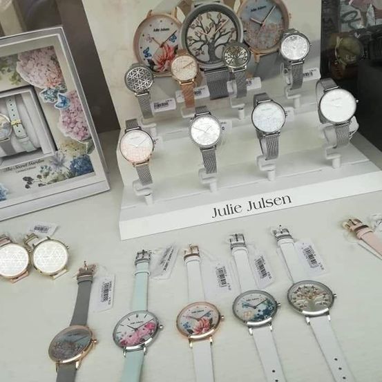 Eine Ausstellung von Julie Julsen Uhren mit verschiedenen Designs und Farben, auf einem weißen Tisch angeordnet. Im Hintergrund ein gerahmtes Bild mit Blumenbildern.