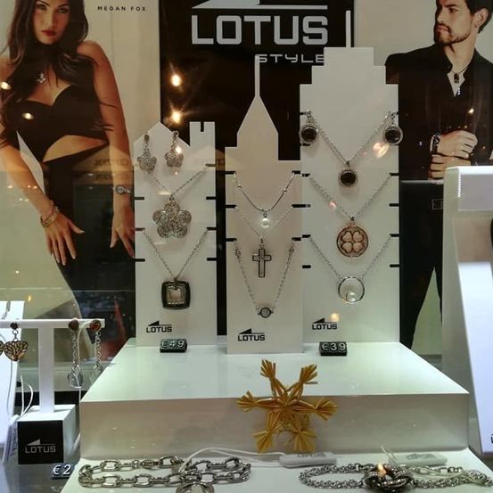Eine Schmuckausstellung mit verschiedenen Halsketten und Ohrringen, gekennzeichnet mit Lotus Style. Die Ausstellung umfasst drei Ständer mit Halsketten zu Preisen von 49, 39 und 2 Euro. Der Hintergrund zeigt ein Modell in einem schwarzen Top und Rock.