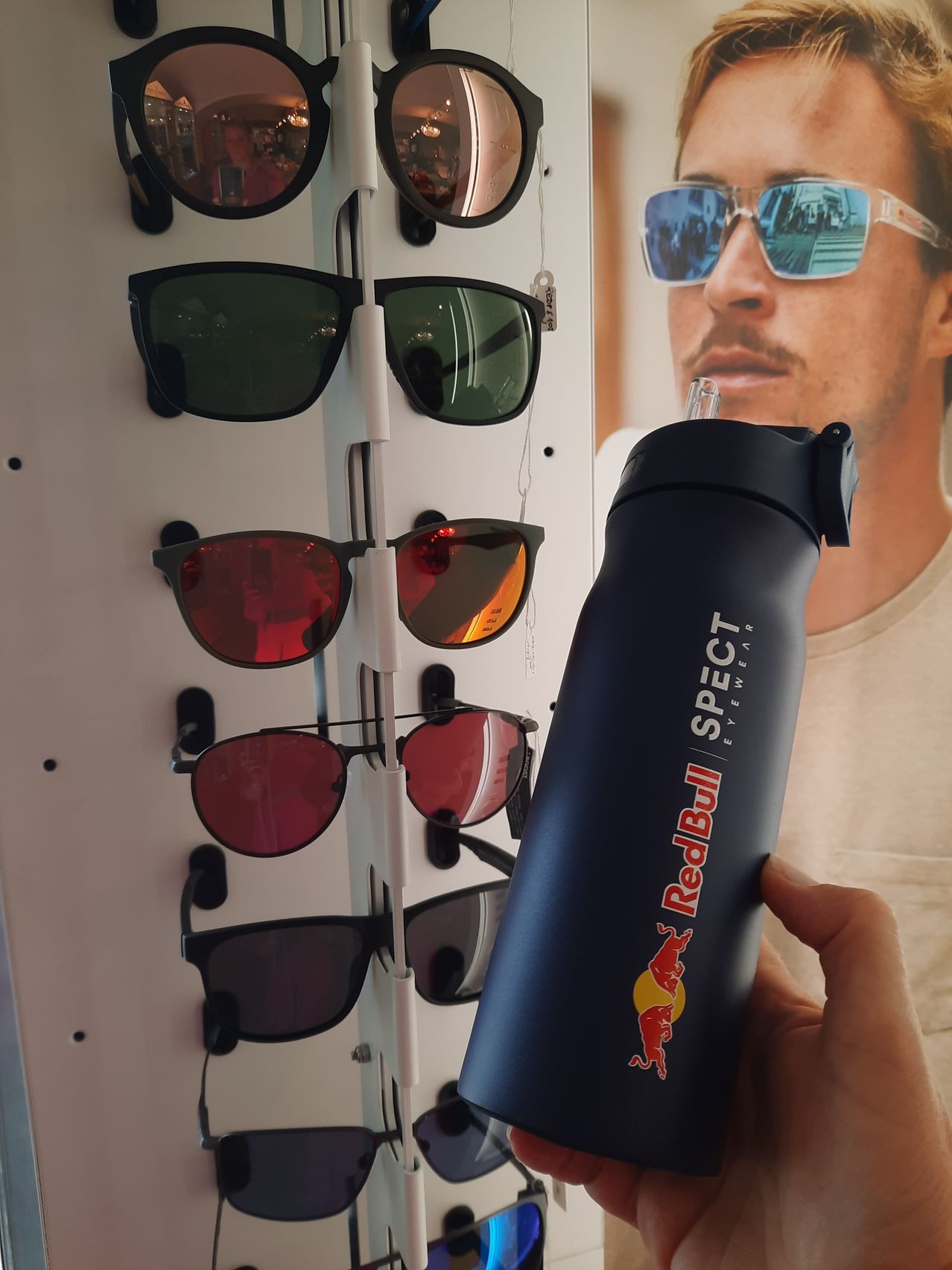 Eine Person hält eine Red Bull Flasche, dahinter sind verschiedene Sonnenbrillen ausgestellt. Die Sonnenbrillen haben verschiedene Farben und Stile.