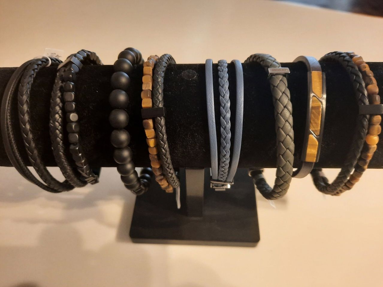 A collection of leather and beaded bracelets are displayed on a black stand. The bracelets include various colors and styles, such as braided, textured, and beaded.
