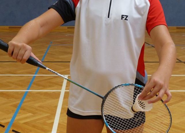 A woman holds a badminton racket on an indoor court, wearing a white and red sports shirt with 'FZ' on the front.