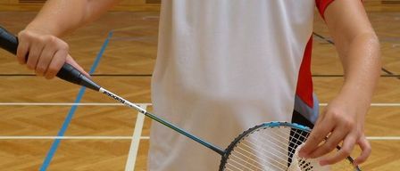 Eine Frau in einem roten, weißen und schwarzen Sporttrikot hält einen Badmintonschläger in der Hand, bereit zum Spielen auf einem Platz mit blauen Markierungen.