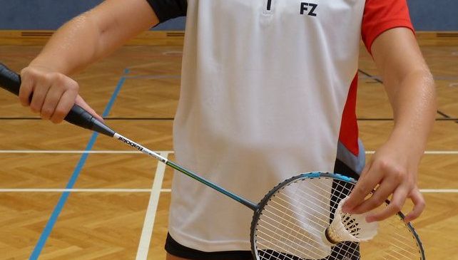 Eine Frau in einem weißen und roten Badminton-Trikot hält einen Badmintonschläger und ist bereit zum Aufschlag auf einer Halle.