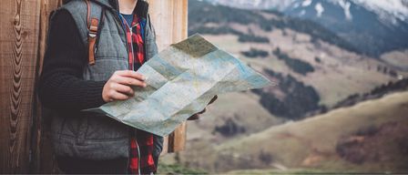 Eine Person steht auf einem Berg, hält eine Karte in der Hand, trägt einen Rucksack und eine Weste.
